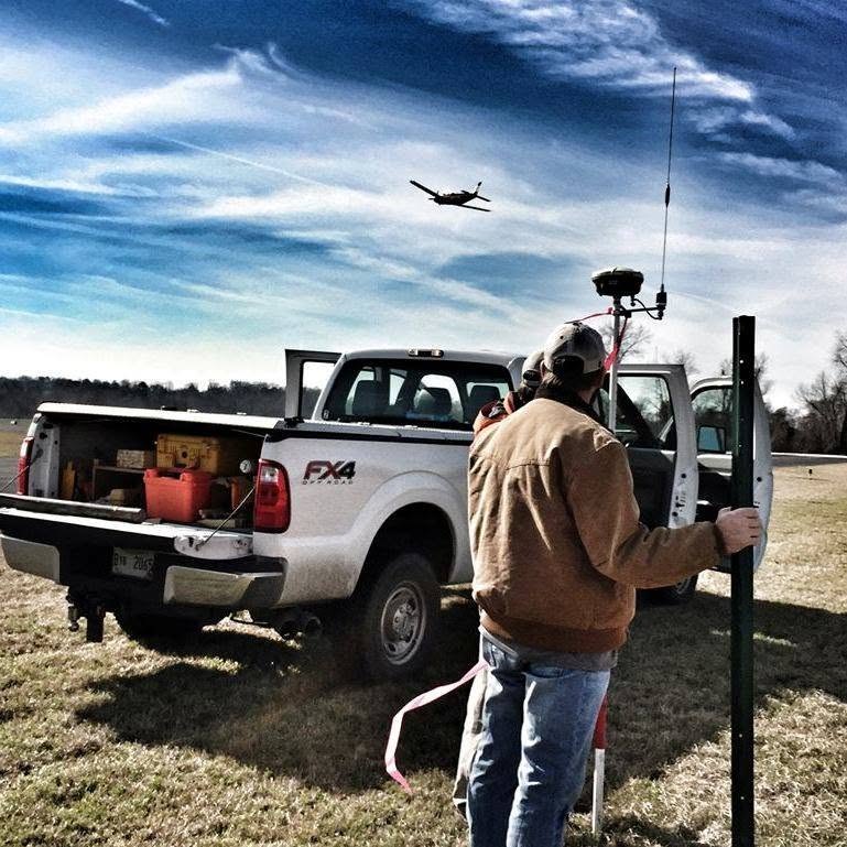 DIMCO surveyor with a GNSS receiver at a field truck while a survey aircraft passes overhead